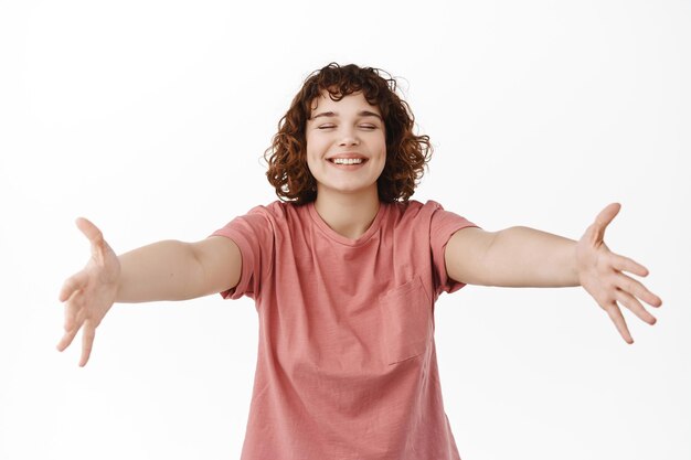 Portret van vrolijke jonge vrouw spreidde handen zijwaarts, reikend naar knuffels en warm welkom, gelukkig en vriendelijk glimlachend naar de camera, staande tegen een witte achtergrond