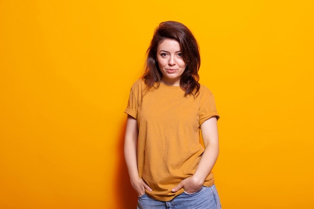 Portret van vrolijke persoon poseren met vertrouwen, hand in hand in jeans zakken. Blanke vrouw die zich positief voelt en over een oranje achtergrond staat, kijkend naar de camera. Zelfverzekerde volwassene