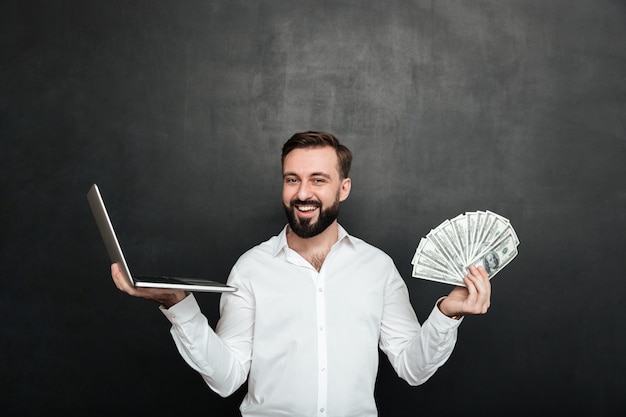 Portret van vrolijke rijke man in wit shirt winnen veel geld dollar valuta met zijn notitieblok over donkergrijs