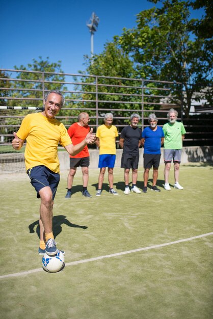 Portret van vrolijke senior man op voetbalveld. Teamleider met grijs haar in sportkleding die staat, bal schopt, teamgenoten op de achtergrond. Voetbal, sport, vrije tijd concept