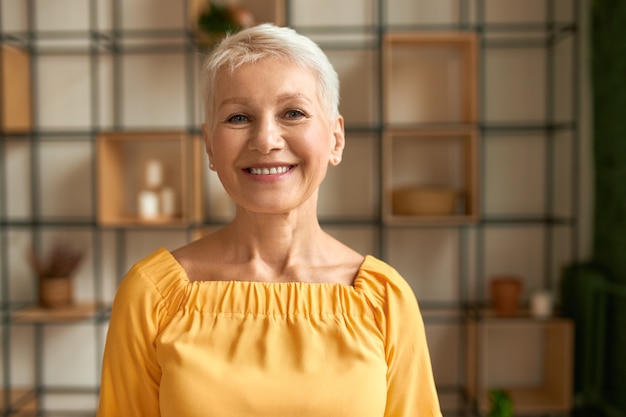 Portret van vrolijke stijlvolle vrouw van middelbare leeftijd met kort kapsel poseren binnenshuis positieve emoties te uiten