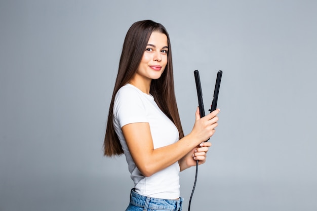 Portret van vrolijke vrouw die gelijkrichter voor haar krullend haar gebruiken die voor de vakantie comfortabel gemakkelijk kapsel van de gebeurtenisdatum voorbereidingen treffen dat op grijs wordt geïsoleerd