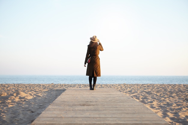 Portret van vrouw het lopen op promenade die blauwe overzees bekijkt die klassieke hoofddeksel en bruine laag draagt