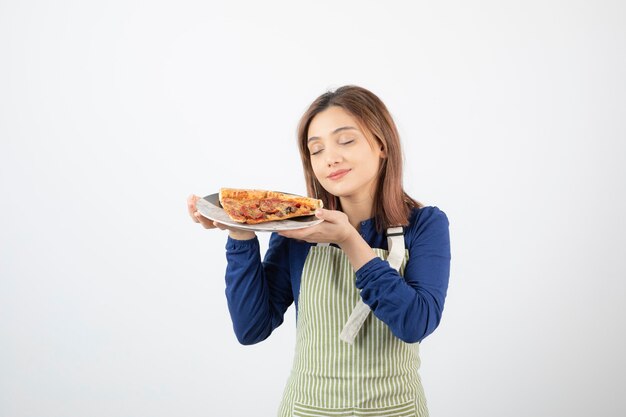 Portret van vrouw in schort ruikt stuk pizza op wit