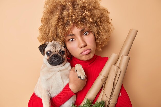 Portret van vrouw met krullend haar houdt handwerk kerstkrans houdt papierrollers poses pug dog portemonnees lippen ziet er helaas rode coltrui geïsoleerd over beige achtergrond. Vakantie concept