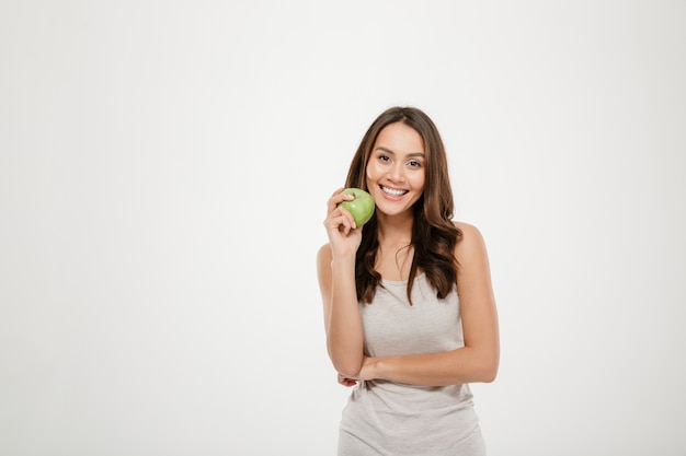 Portret van vrouw met lang bruin haar die op camera met groene in hand appel kijken, die over wit wordt geïsoleerd