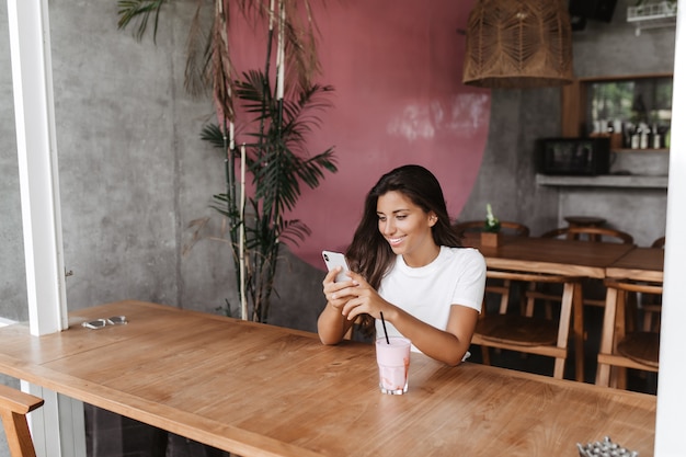 Portret van vrouw zitten in café en chatten met glimlach in smartphone