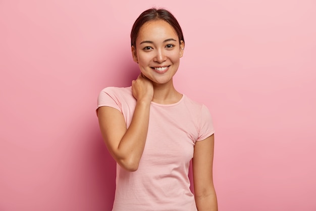 Portret van vrouwelijk meisje met aangename glimlach, zachte blik, nek aanraakt, gezichtsuitdrukking verheugd heeft, aangenaam gesprek heeft met goede vriend, gekleed in casual outfit, geïsoleerd op roze muur