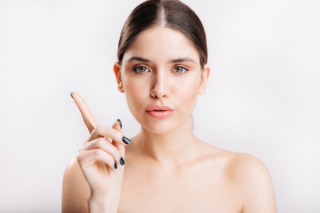 Portret van vrouwelijk model zonder make-up die met wijsvinger naar omhoog op geïsoleerde muur richt.