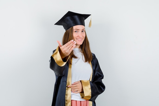 Portret van vrouwelijke afgestudeerde die uitnodigt om in academische kleding te komen en vrolijk vooraanzicht kijkt