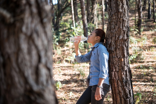 Portret van vrouwelijke backpacker drinkt zoet water uit de fles terwijl hij rugzak in het dennenbos draagt.
