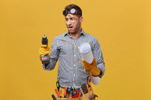 Portret van vuile reparateur met gereedschapsriem met blauwdruk en boormachine die ernaar kijkt met woede omdat het slecht werkt. Ambachtsman uiting van zijn ongenoegen over gereedschappen en instrumenten