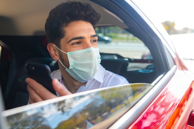 Portret van zakenman die gezichtsmasker draagt en zijn mobiele telefoon gebruikt op weg om in een auto te werken. Bedrijfsconcept. Nieuw normaal levensstijlconcept.