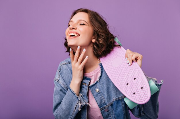 Portret van zalig wit meisje met golvend kapsel. schitterende vrouw die met skateboard met gesloten ogen glimlacht.