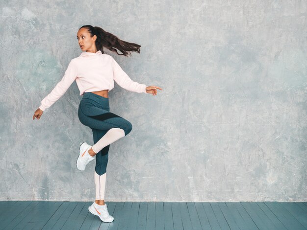 Portret van zekere fitness vrouw in sporten kleding die zeker kijken. Vrouwelijk springen in studio dichtbij grijze muur