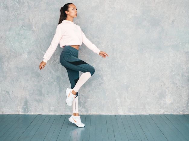 Portret van zekere fitness vrouw in sporten kleding die zeker kijken. Vrouwelijk springen in studio dichtbij grijze muur