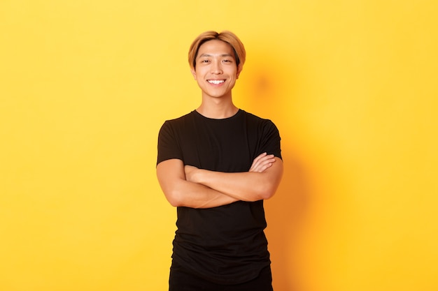Portret van zelfverzekerde knappe Aziatische man tevreden glimlachend, staande over gele muur in zwarte kleding.