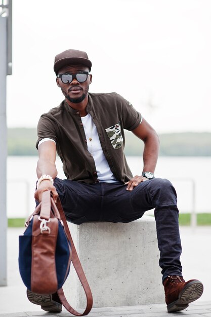 Portret van zittende stijlvolle Afro-Amerikaanse man draagt een zonnebril en pet buiten Street fashion zwarte man