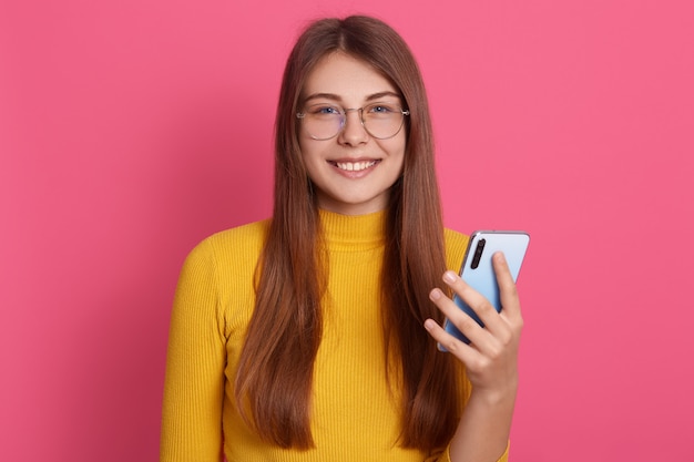 Portret van zoete oprechte aanbiddelijke aantrekkelijke jonge vrouw met een aangename glimlach, met smartphone, in een goed humeur, permanent geïsoleerd over roze muur. mensen en technologie concept.