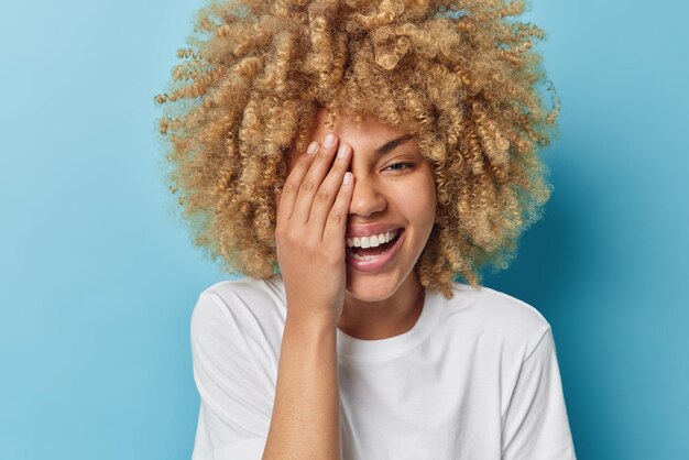 Portret van zorgeloze jonge mooie vrouw met krullend haar bedekt de helft van het gezicht met palm lacht graag uit gekleed in casual wit t-shirt geïsoleerd op blauwe achtergrond Gelukkig emoties concept