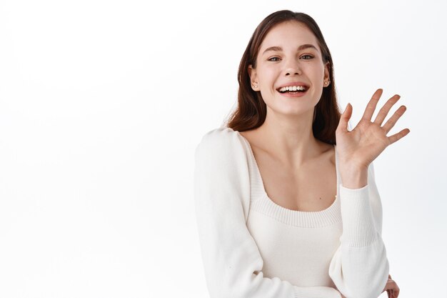 Portret van zorgeloze vriendelijke Europese vrouwelijke ondernemer, hand op de borst en breed glimlachend terwijl ze met opgeheven hand zwaait, vriend groet, ontmoeting met vrienden van het werk in café