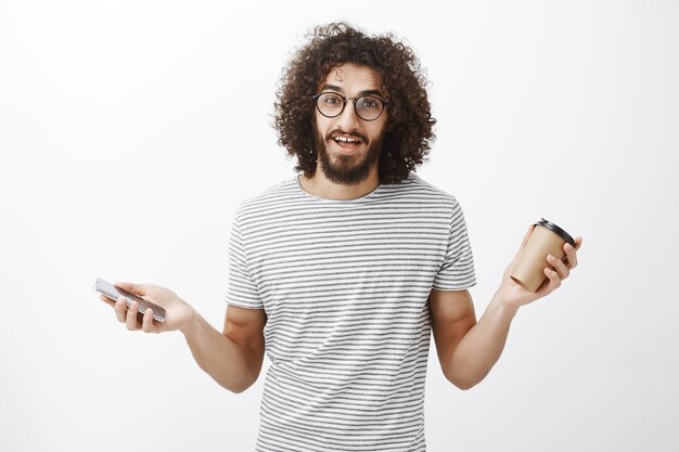 Portret van zorgeloze vriendelijke knappe man in glazen en gestreept shirt, handen spreiden met kopje koffie en smartphone, gepassioneerd praten over nieuw spel