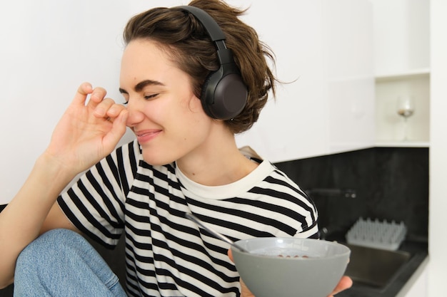 Portret van zorgeloze vrouw met koptelefoon die haar ontbijt eet met een kom ontbijtgranen in de hand en lacht