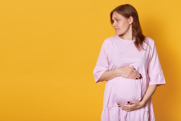 Portret van zwangere vrouw met bruin haar, draagt roze poeder jurk, model staat geïsoleerd op geel, kijkt opzij, raakt buik. Kopieer ruimte voor promotionele tekst of reclame