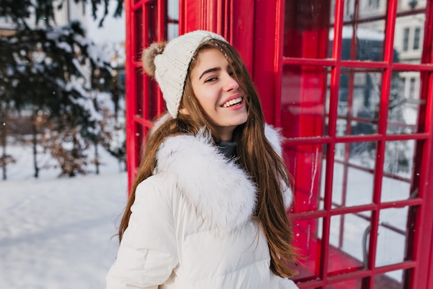 Portret vrolijke jonge vrouw in warme gebreide muts met lang donkerbruin haar genieten van bevroren winterweer op straat op rode telefooncel