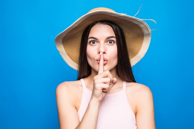 Portretclose-up van vrouw die strohoed draagt die vinger toont aan lippen om geheim over blauwe muur te houden.