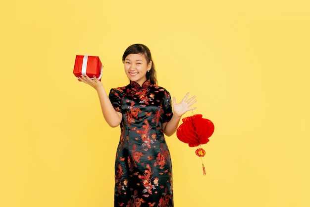 Poseren met lantaarn en cadeau, glimlachend. Gelukkig Chinees nieuwjaar. Aziatisch jong meisje portret op gele achtergrond. Vrouwelijk model in traditionele kleding ziet er gelukkig uit. Copyspace.