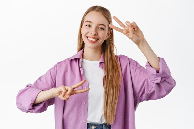 Positief blond meisje, glimlachend en vrede v-teken gebaren tonend, kawaii kijkend naar de camera, zich gelukkig voelend terwijl ze tegen een witte achtergrond staat