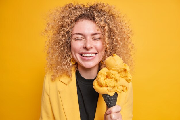 Positief duizendjarig meisje met krullend borstelig haar glimlacht gelukkig heeft plezier met zomerijs geniet van vakantie heeft een vrolijke stemming gekleed in formele kleding geïsoleerd over gele muur. Lekker dessert