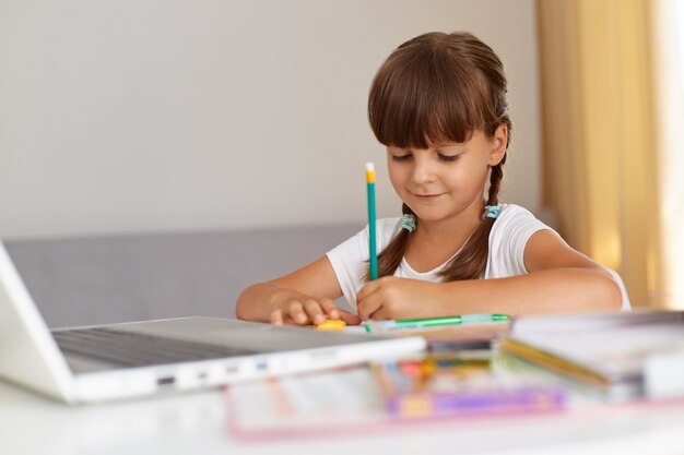 Positief, goed uitziend schoolmeisje dat casual kleding draagt, in oefenboek schrijft, een positieve stemming heeft, aan tafel zit in de woonkamer, online onderwijs.