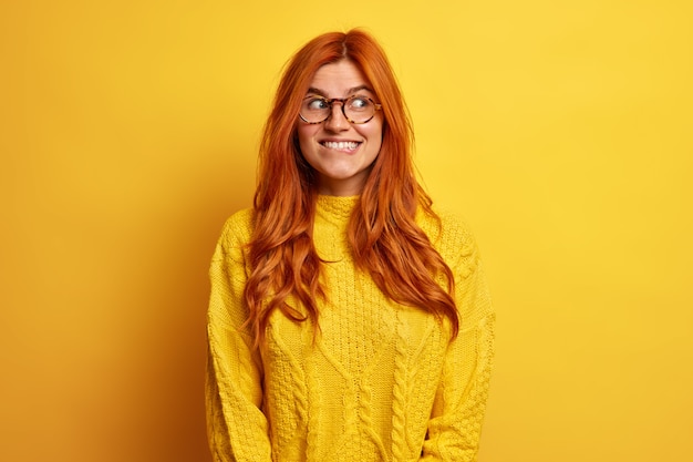 Positief goed uitziende jonge vrouw heeft natuurlijke roodharige looks met een vrolijke mysterieuze uitdrukking opzij bijt lippen nadenkt over mooi aanbod gekleed in casual trui.