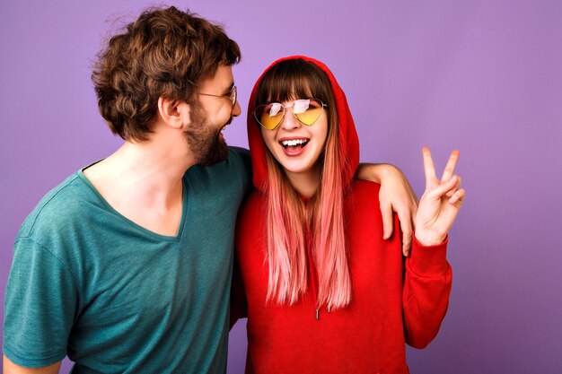 positief grappig portret van een gelukkig stel dat samen plezier heeft, knuffels en lachen, familie en liefde, casual jeugdkleren en accessoires, vredesgebaar, violette muur, relatie