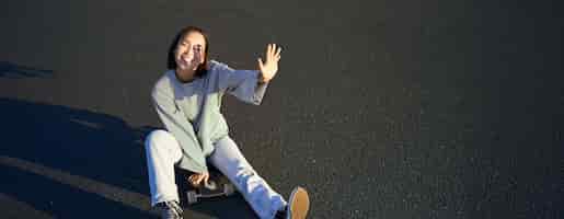 Gratis foto positief koreaans meisje bedekt haar gezicht tegen het zonlicht zit op skateboard en glimlacht gelukkig