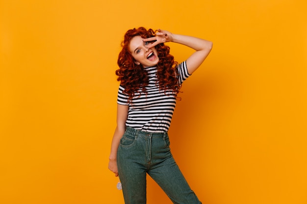 Positief meisje met golvend rood haar dat lolly houdt en emotioneel camera bekijkt, vredesteken toont.