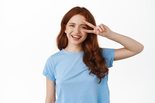 Positief roodharig krullend meisje toont vredesv-teken en glimlacht breed, staat ontspannen in casual kleding met vrolijke gezichtsuitdrukking, witte achtergrond