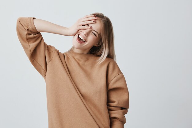 Positief schattig vrouwelijk model poseert, heeft een stralende aangename glimlach, is blij om complimenten te ontvangen, verheugt zich in de vrije tijd. Glimlachende vrolijke blondevrouw die binnen ontspannen.