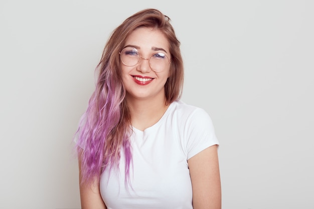 Positief tevreden jong meisje in glazen en wit t-shirt met gelukkige brede glimlach, die in uitstekend humeur is, geïsoleerd over grijze muur poseren.