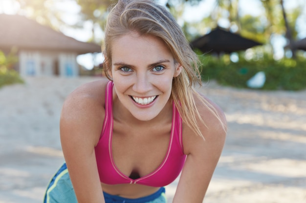 Positieve aangenaam uitziende lachende jonge vrouw gekleed in actieve slijtage, geniet van sportieve levensstijl, treinen op het strand tijdens zonnig zomerweer. Vrouwelijke atleet doet actieve sportoefeningen buiten.