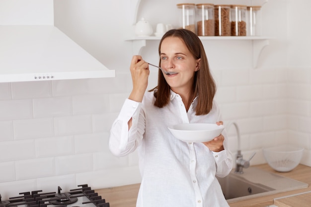 Positieve aantrekkelijke jonge volwassen vrouw met donker haar die een wit casual overhemd draagt. ontbijten, bord in handen houden, wegkijken, van lepel eten.