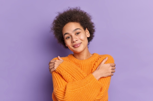 Positieve Afro-Amerikaanse tienermeisje knuffelt zichzelf raakt schouders spreekt zelfliefde glimlacht aangenaam gekleed in gebreide oranje trui.