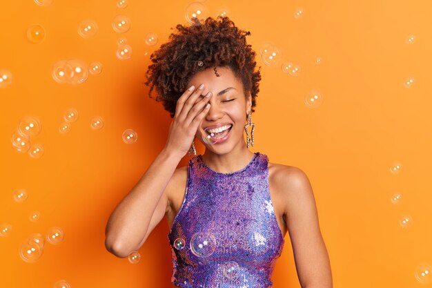 Positieve Afro-Amerikaanse vrouw heeft betrekking op gezicht met handglimlach in het algemeen heeft perfecte witte tanden gekleed in stijlvolle kleding geïsoleerd over oranje muur met zeepbellen rond