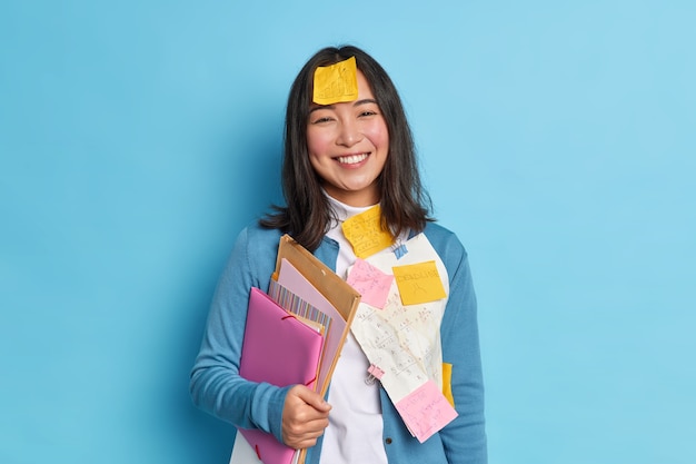 Positieve Aziatische student houdt mappen staat blij met stickers op kleding en voorhoofd bereidt projectwerk in de economie voor, graag klaar met het doen van belangrijke taak draagt trui.