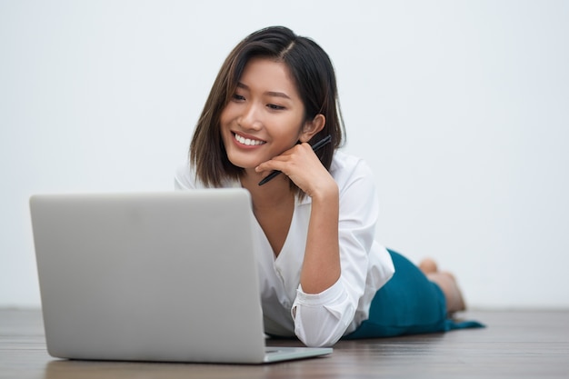 Positieve Aziatische vrouw liggend op de vloer met laptop
