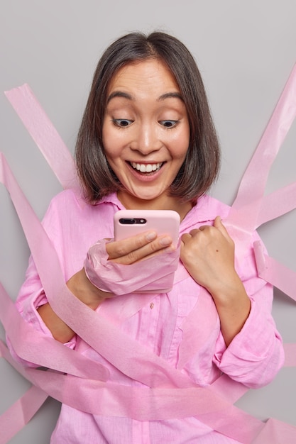 Positieve aziatische vrouw met blije uitdrukking controleert melding op smartphone controleert evenementen die in de buurt gaan en bekijkt geweldig nieuws dat met tapes aan de muur is gepleisterd, kijkt naar brievenbustypen op het scherm van de mobiele telefoon