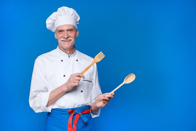 Positieve bejaarde chef-kok met houten lepel en vork, in wit uniform en pet poseren geïsoleerd op blauwe muur