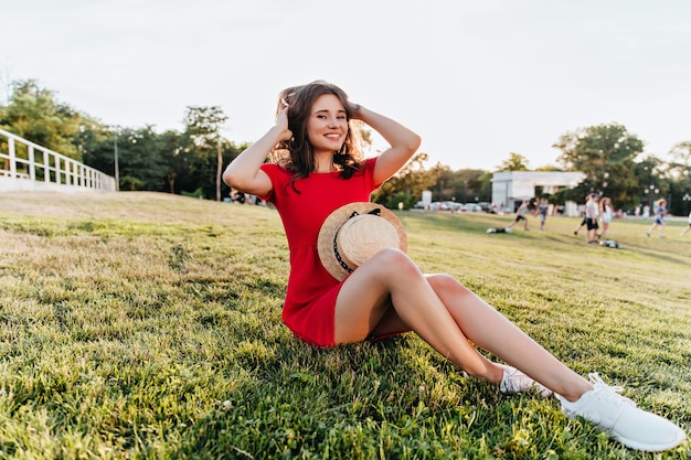 Positieve blanke meisje zittend op het gras en spelen met haar bruine haar. Vrolijke lachende dame in rode jurk genieten van weekendochtend in park.